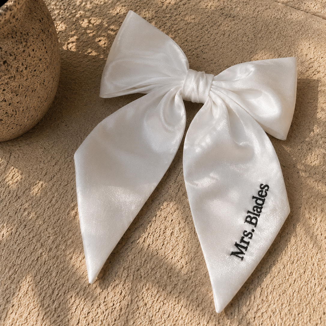 A white satin bow hair accessory with the text 'Mrs. Blades' embroidered in black on one loop, displayed on a textured beige stone surface next to a potted plant.