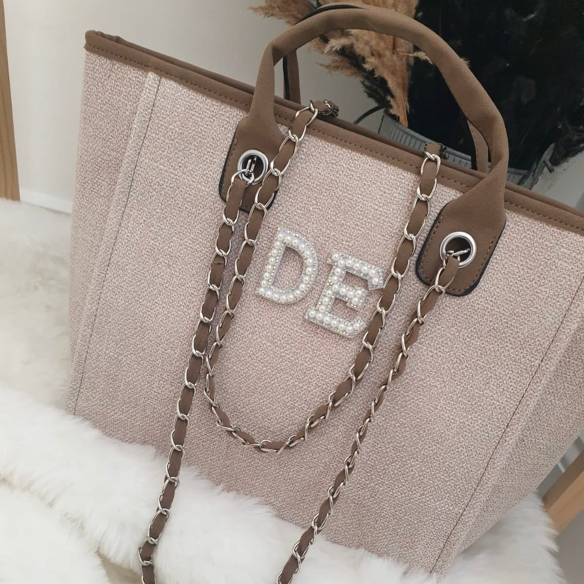 Beige handbag with brown chain strap and 'DE' initials on a white surface.