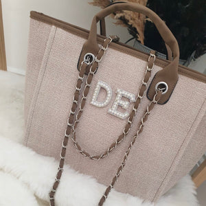 Beige handbag with brown chain strap and 'DE' initials on a white surface.