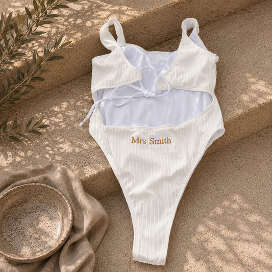 A white ribbed one-piece swimsuit with a cutout back, tie detail, and 'Mrs Smith' embroidered in gold on the front, laid flat on beige stone steps with a bowl and greenery nearby.