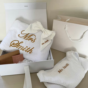White towel and bikini with 'Mrs Smith' embroidery in a gift box on a wooden surface.