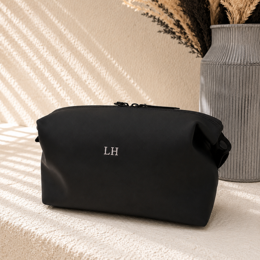 A black zippered cosmetic pouch with silver 'LH' initials on the front, styled on a textured cream surface with a gray ribbed vase in the background.
