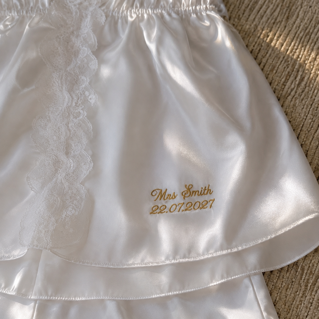 White satin pajama shorts with a lace side panel and personalized gold embroidery reading 'Mrs. Smith 22.