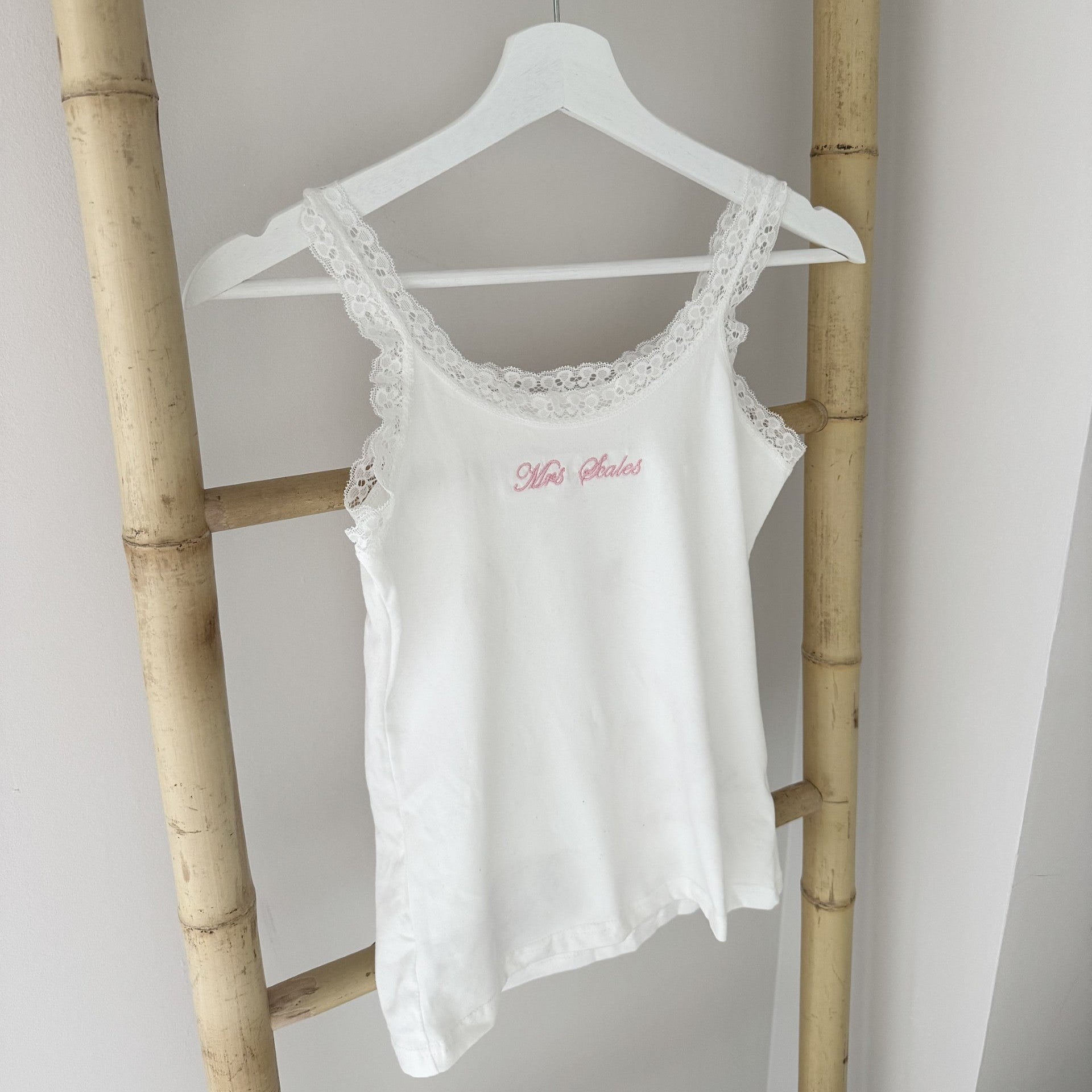White lace-trimmed tank top with pink text on a hanger against a light gray wall.