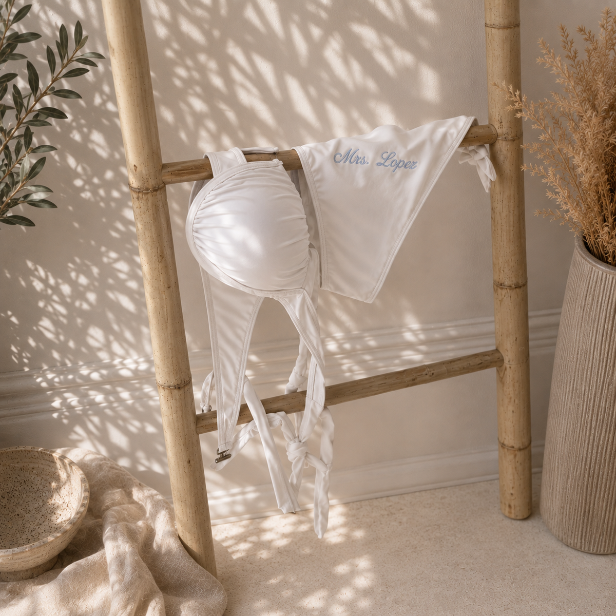 A white string bikini top with a matching white sarong-style bottom draped over a bamboo stand in a styled interior setting with warm sunlight and leaf shadows.