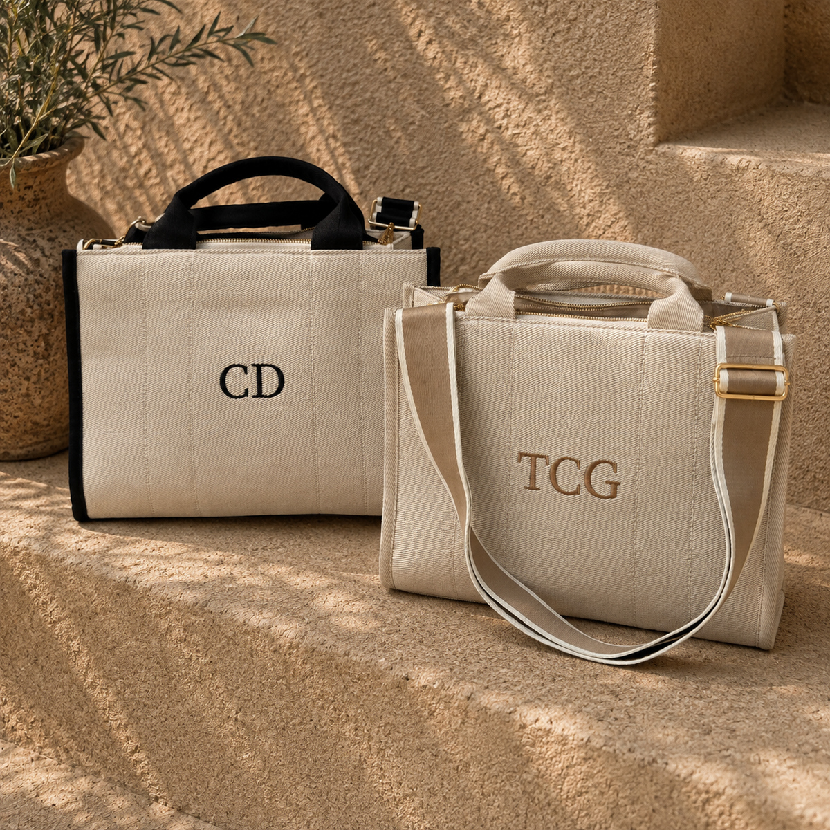 Two structured canvas tote bags in warm beige tones, styled on sunlit stone steps. One bag has black trim and handles with the monogram 'CD' in black, and the other has beige trim and handles with 'TCG' in gold-toned embroidery.