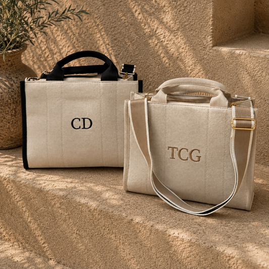 Two structured canvas tote bags in warm beige tones, styled on sunlit stone steps. One bag has black trim and handles with the monogram 'CD' in black, and the other has beige trim and handles with 'TCG' in gold-toned embroidery.