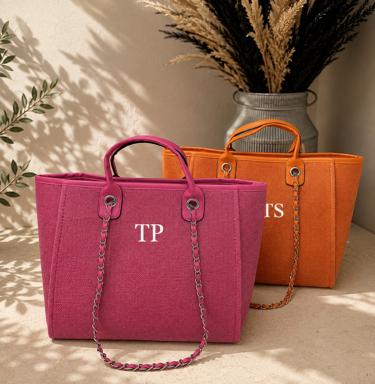 Two canvas tote bags in magenta pink and orange, styled on a beige surface with a neutral background and a vase of dried grass. The front bag is magenta pink with a pink and silver chain strap and a white monogram 'TP' on the front.