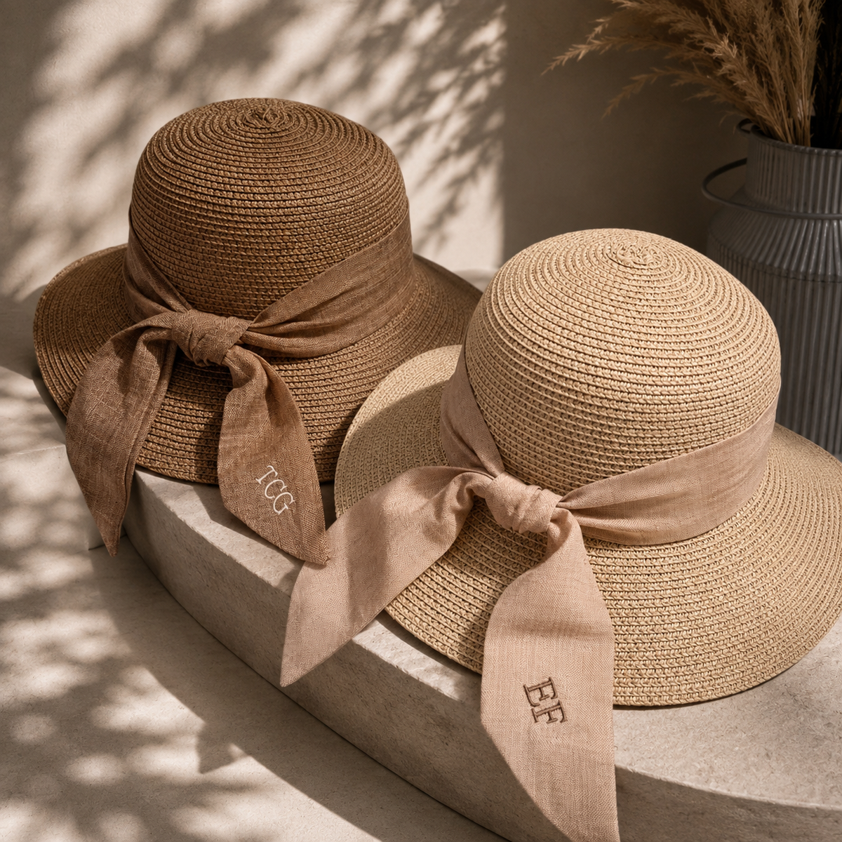 Two woven straw sun hats with wide brims and ribbon bows, styled on a stone surface in warm natural light. One hat is tan with a darker brown ribbon and embroidered 'TCG' text, and the other is light beige with a tan ribbon and embroidered 'EF´ text.