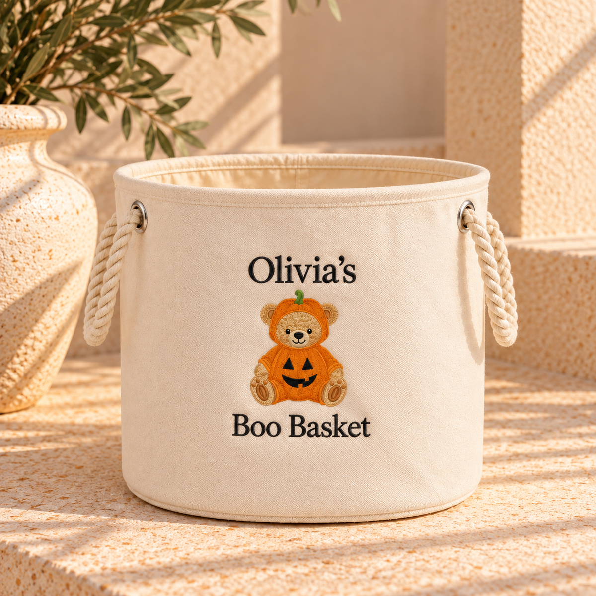A cream-colored fabric storage basket with rope handles, personalized with the text 'Olivia's Boo Basket' and a cartoon teddy bear in a pumpkin costume. The basket is styled on a textured stone surface next to a ceramic vase with green leaves.