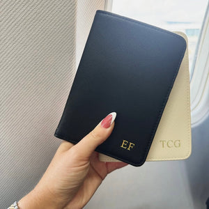 Hand holding a black passport holder with gold initials against a neutral background