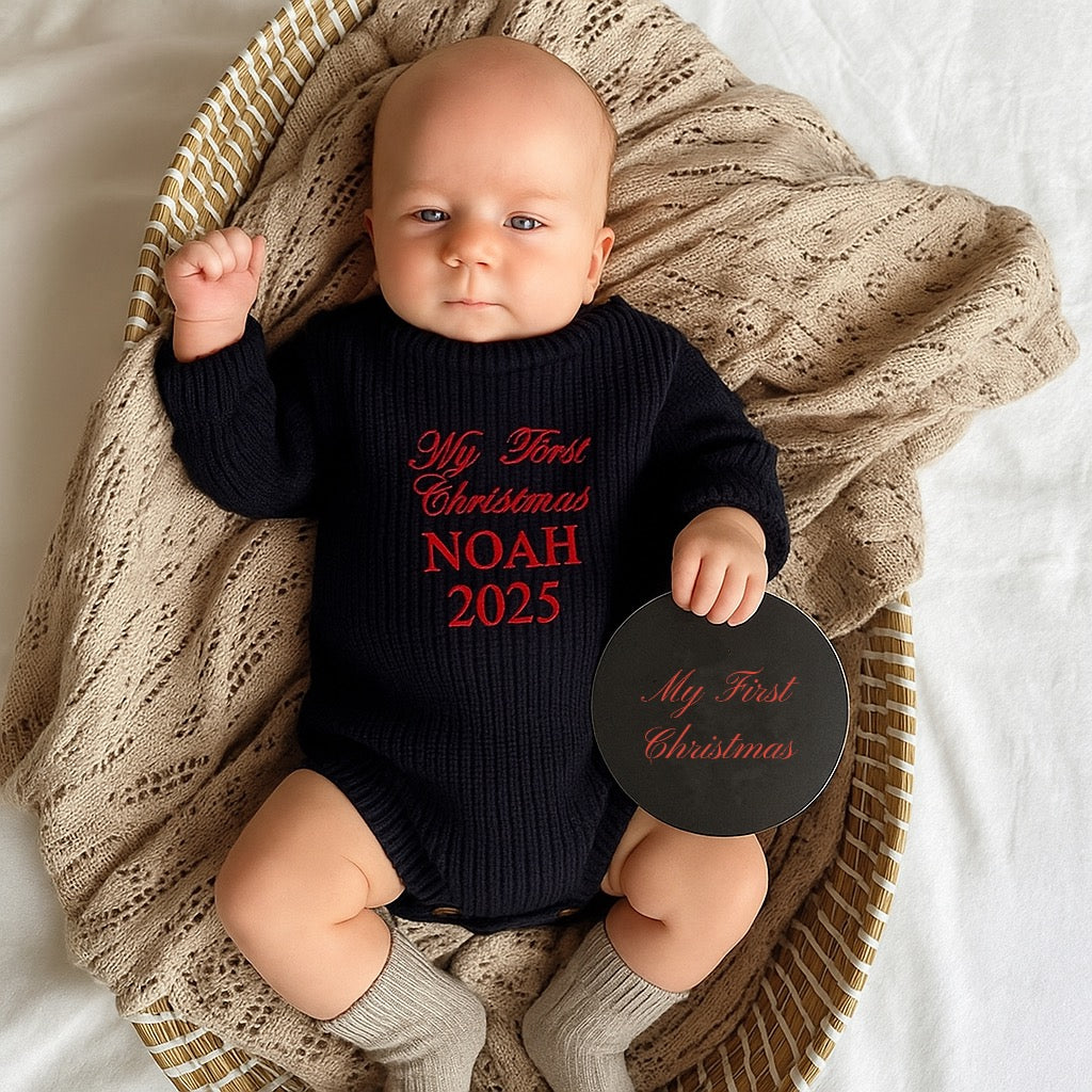 Baby wearing a 'My First Christmas NOAH 2025' onesie, holding a disc with the same text, sitting in a woven basket.