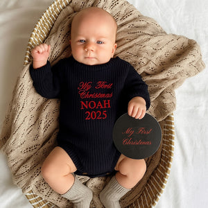 Baby wearing a 'My First Christmas NOAH 2025' onesie, holding a disc with the same text, sitting in a woven basket.