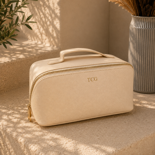 A cream-colored faux leather cosmetic bag with a top handle, gold zipper, and 'TCG' logo, styled on a stone surface with warm sunlight and shadows.