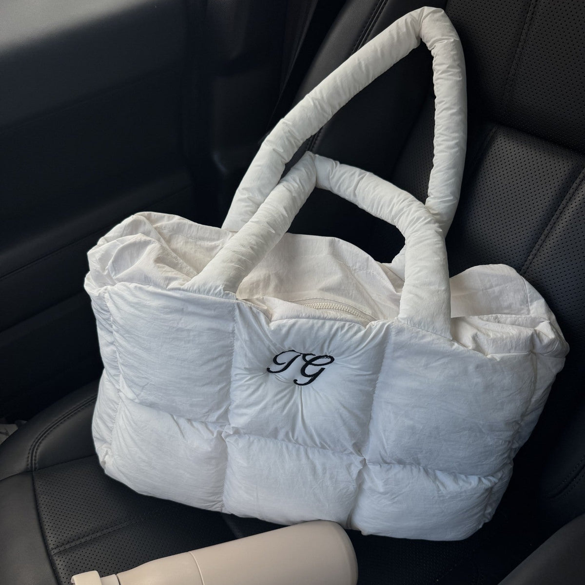 A white puffer tote bag with thick padded handles and a black monogram logo, resting on a black car seat next to a beige water bottle and keys.