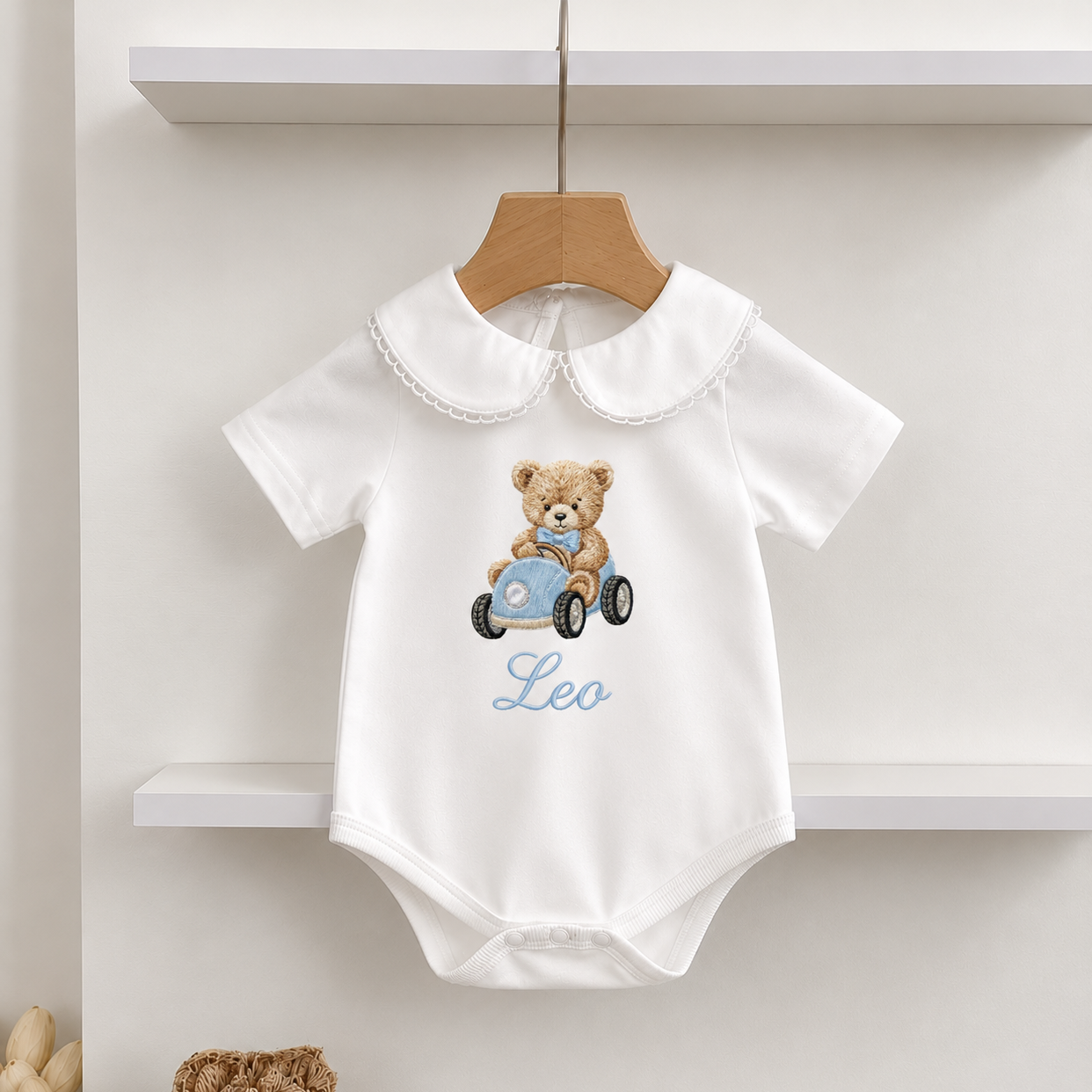 A white short-sleeve baby bodysuit with a Peter Pan collar, featuring an embroidered teddy bear driving a blue car and the name 'Leo' in light blue script. The bodysuit is displayed on a wooden hanger hanging from a white shelf.