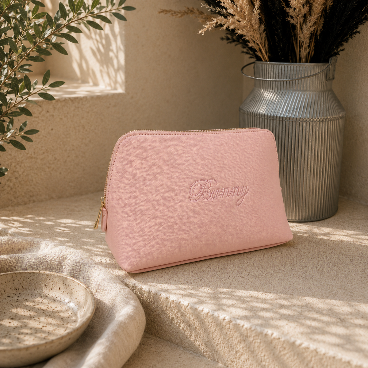 A dusty pink zippered cosmetic pouch with the name 'Bunny' embossed on the front, styled on a beige stone surface with a galvanized metal vase and dried grass in the background.