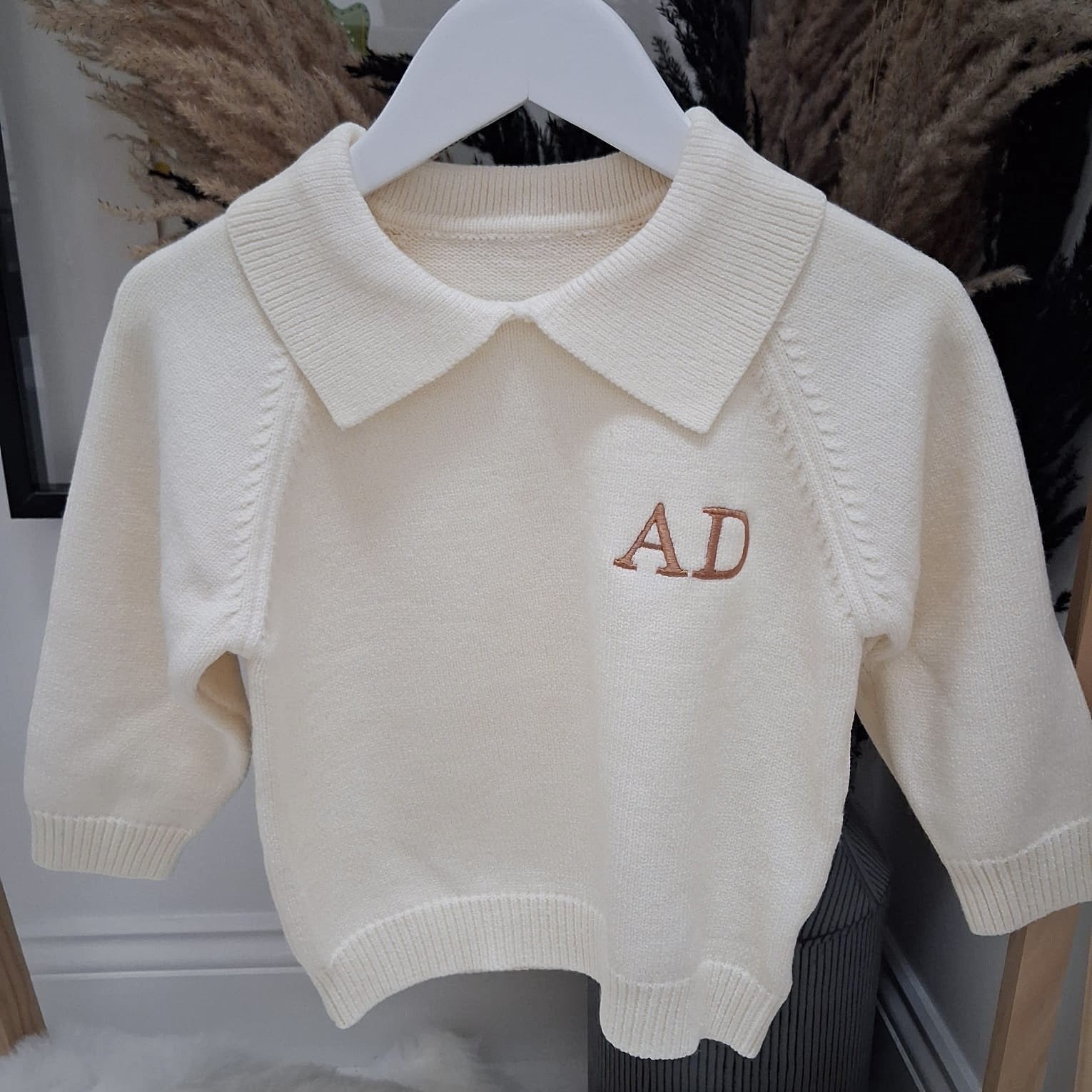Beige sweater with 'AD' embroidery on a hanger against a neutral background