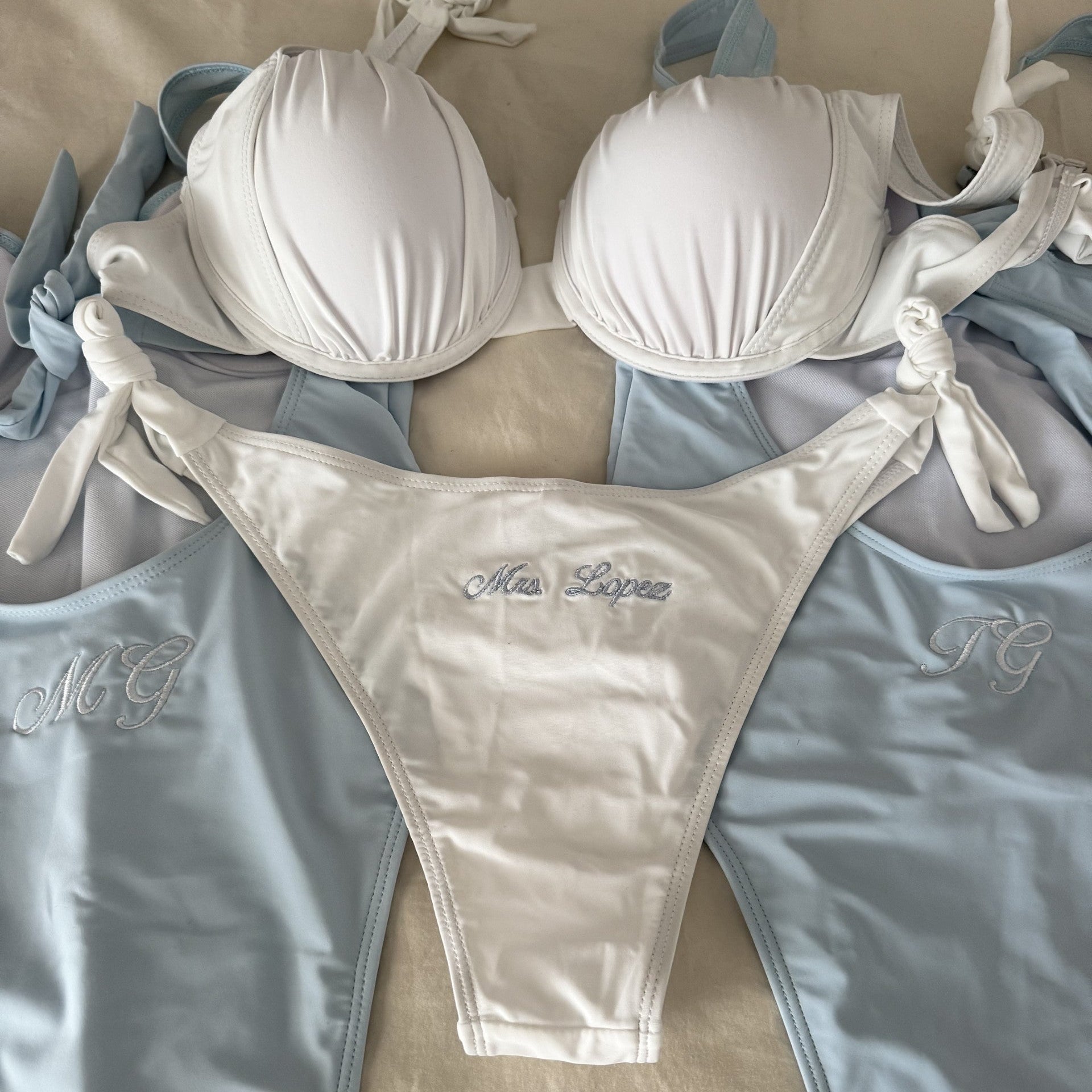 White bikini set with 'Mrs Lopez' embroidery on a beige surface