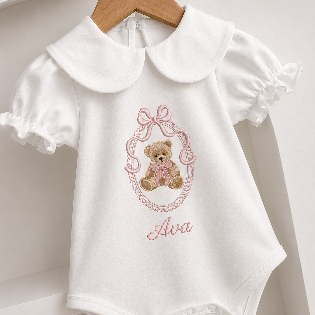 White baby bodysuit with a large Peter Pan collar, short ruffled sleeves, and an embroidered teddy bear design with the name 'Ava' in pink script, hanging on a wooden hanger.