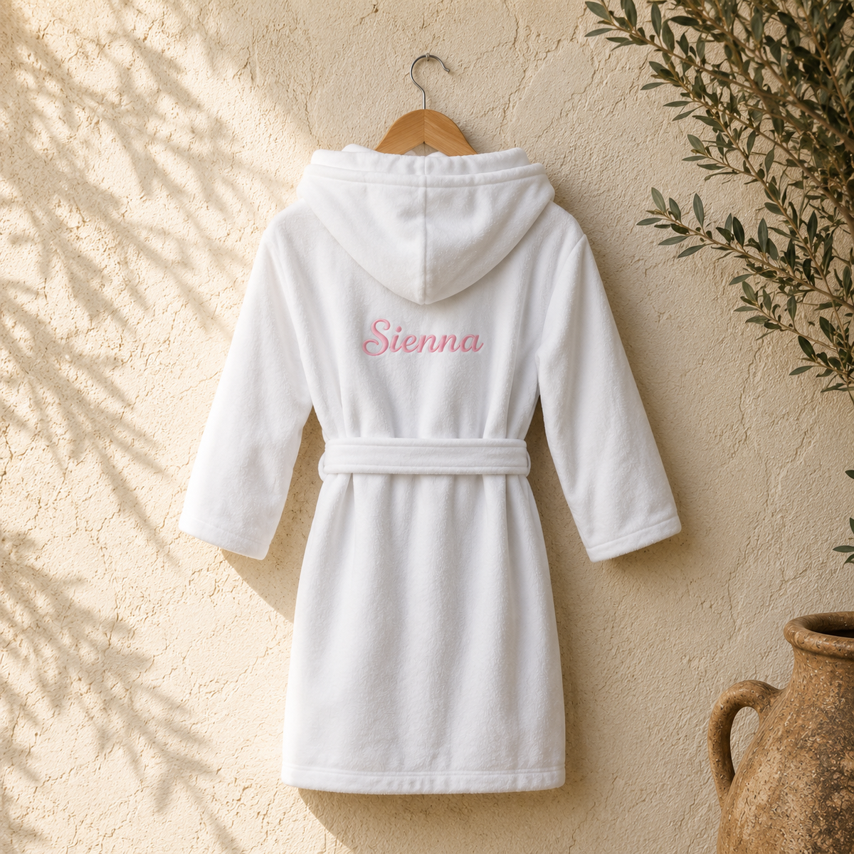 A white hooded bathrobe with a matching waist tie hangs on a wooden hanger against a textured beige wall. The back of the robe is personalized with the name 'Sienna' in pink script embroidery.