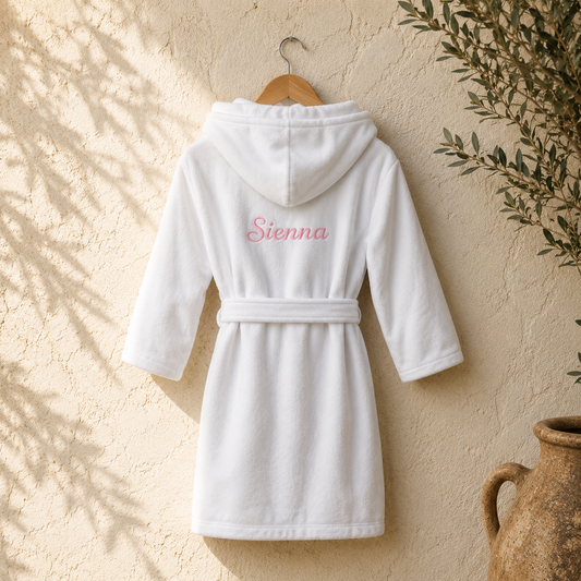 A white hooded bathrobe with a matching waist tie hangs on a wooden hanger against a textured beige wall. The back of the robe is personalized with the name 'Sienna' in pink script embroidery.