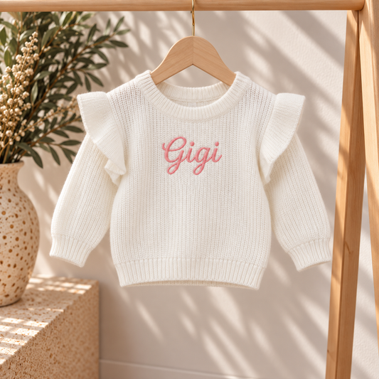 A white ribbed knit baby sweater with ruffled sleeves and the name 'Gigi' embroidered in pink script on the chest, hanging on a wooden hanger on a clothing rack.