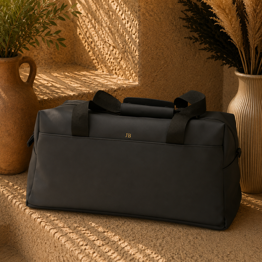 A black duffel bag with top handles sits on a textured stone surface in a warm, sunlit setting with potted plants and dried grass in vases. A small gold 'JB' monogram is visible on the front.