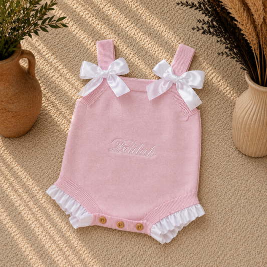 A light pink knit baby romper with white ruffle trim, white bow details on the shoulder straps, and three snap buttons at the crotch. The brand name 'Delilah' is embroidered on the chest.