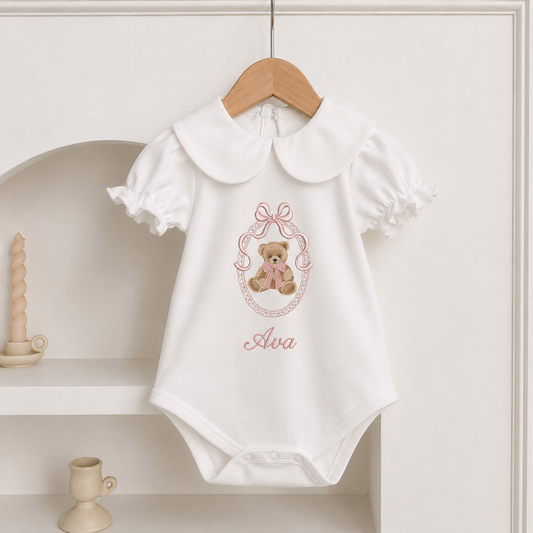 A white short-sleeve baby bodysuit with a large Peter Pan collar, ruffled sleeves, and an embroidered design of a teddy bear in a pink bow frame with the name 'Alva' below, hanging on a wooden hanger against a cream-colored shelf background.
