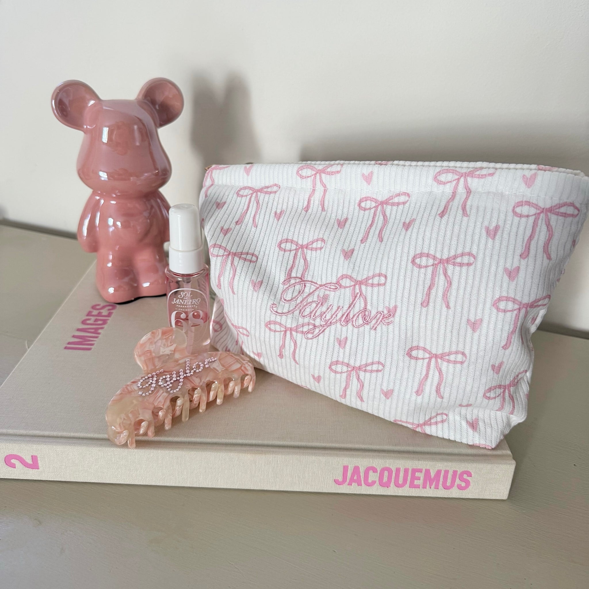 Pink bear figurine, hairbrush, and pouch on a book with 'Jacquemus' branding. with personalised bag