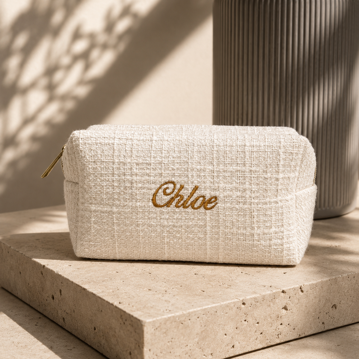 A cream-colored woven cosmetic pouch with gold embroidered 'Chloe' script, resting on a stone pedestal in warm, shadowed sunlight.