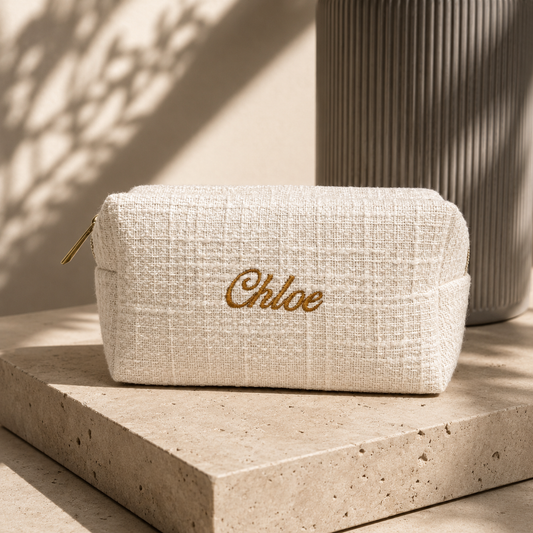 A cream-colored woven cosmetic pouch with gold embroidered 'Chloe' script, resting on a stone pedestal in warm, shadowed sunlight.