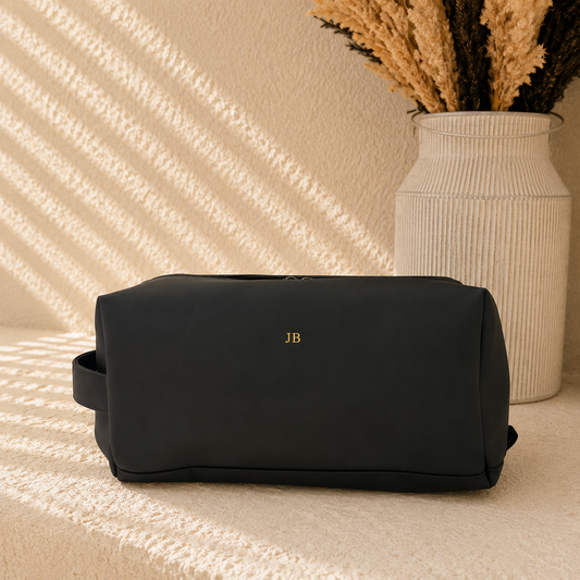 A black rectangular toiletry bag with a small gold 'JB' monogram on the front, styled on a textured beige surface with warm sunlight shadows and a white ribbed vase in the background.