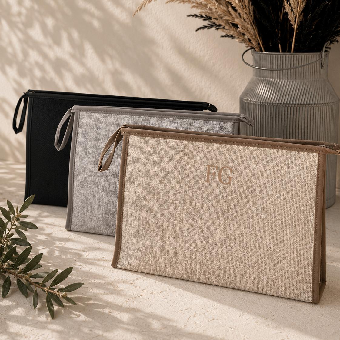 Three woven zip pouches in black, gray, and beige are arranged on a textured surface with dried grass in a metal vase and leafy branches in the background. The front beige pouch has a monogram 'FG' on the front.