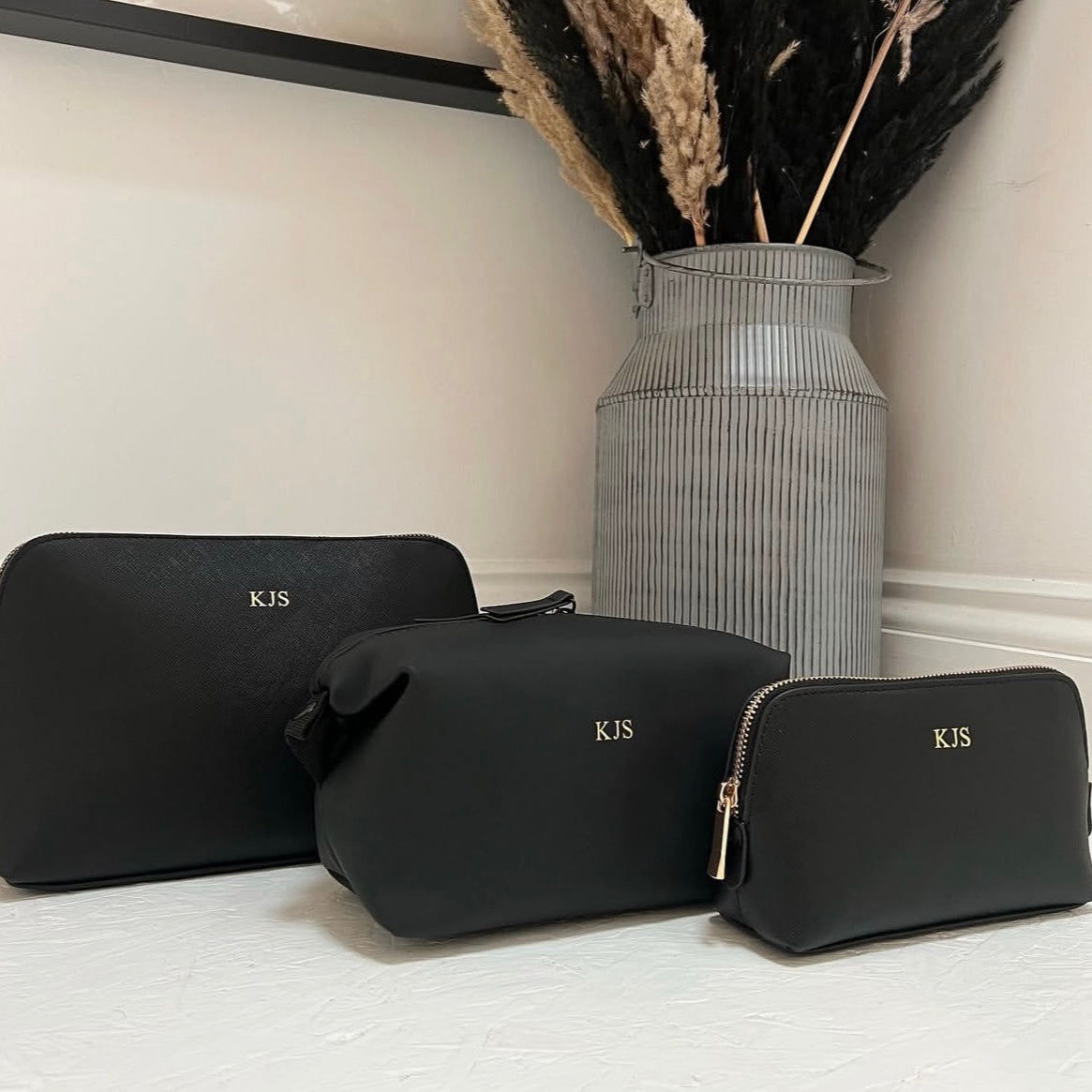 Three black cosmetic bags with 'KJS' monogram on a light surface, with a vase of dried plants in the background.