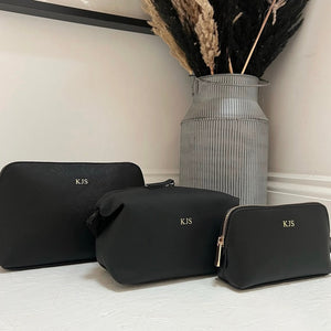 Three black cosmetic bags with 'KJS' monogram on a light surface, with a vase of dried plants in the background.