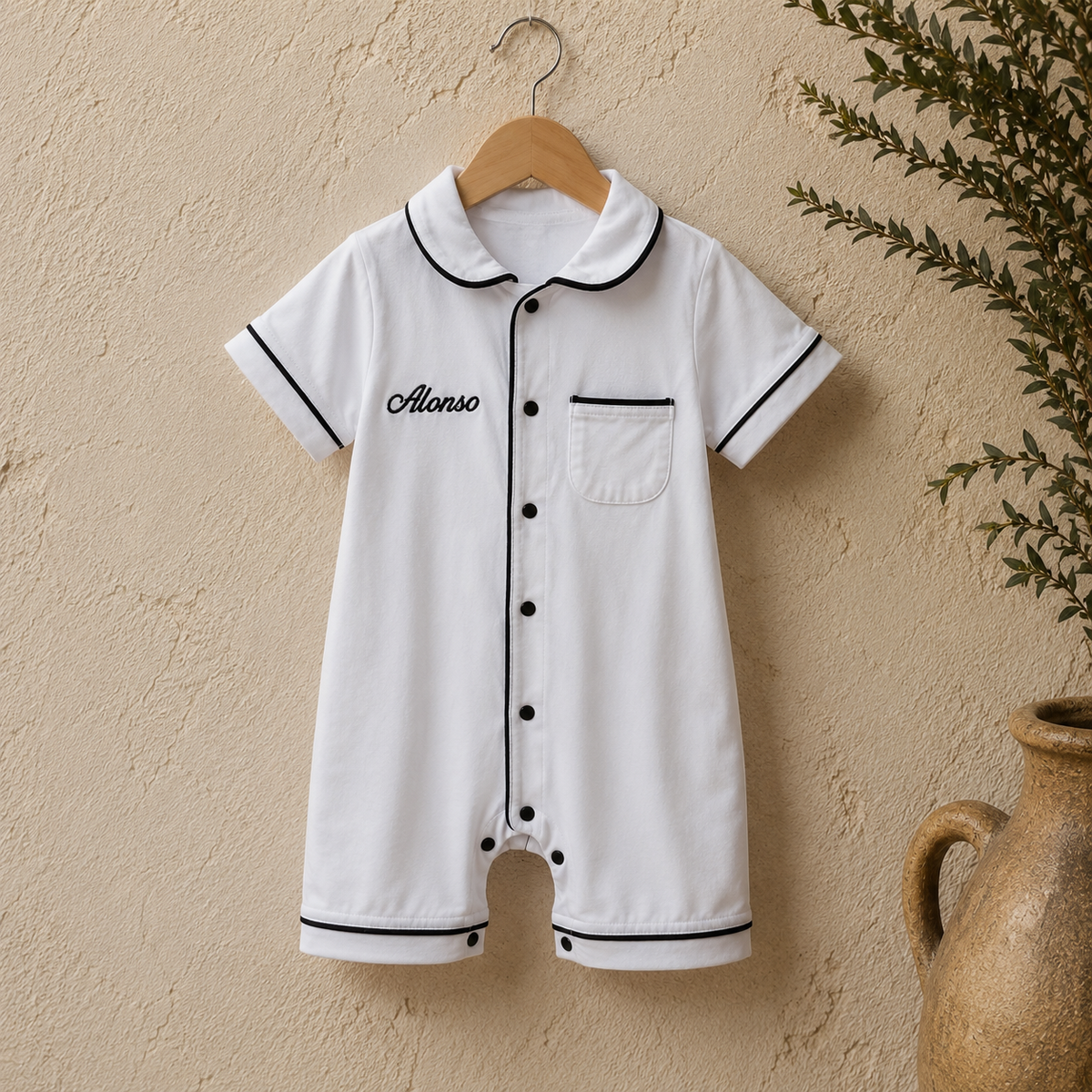 A white short-sleeve baby jumpsuit with black piping and the name 'Alonso' embroidered on the chest, hanging on a wooden hanger against a textured beige wall with a plant and vase in the background.