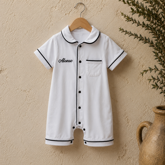 A white short-sleeve baby jumpsuit with black piping and the name 'Alonso' embroidered on the chest, hanging on a wooden hanger against a textured beige wall with a plant and vase in the background.