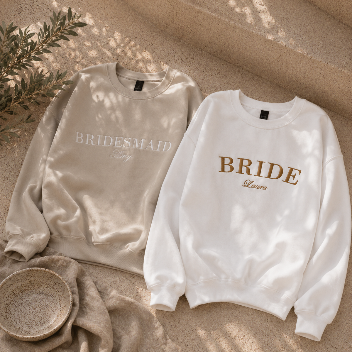 Two crewneck sweatshirts laid flat on a sandy surface with soft shadows. The left sweatshirt is champagne beige with 'BRIDESMAID' in white script, and the right sweatshirt is white with 'BRIDE' in gold and 'Lena' in cursive.