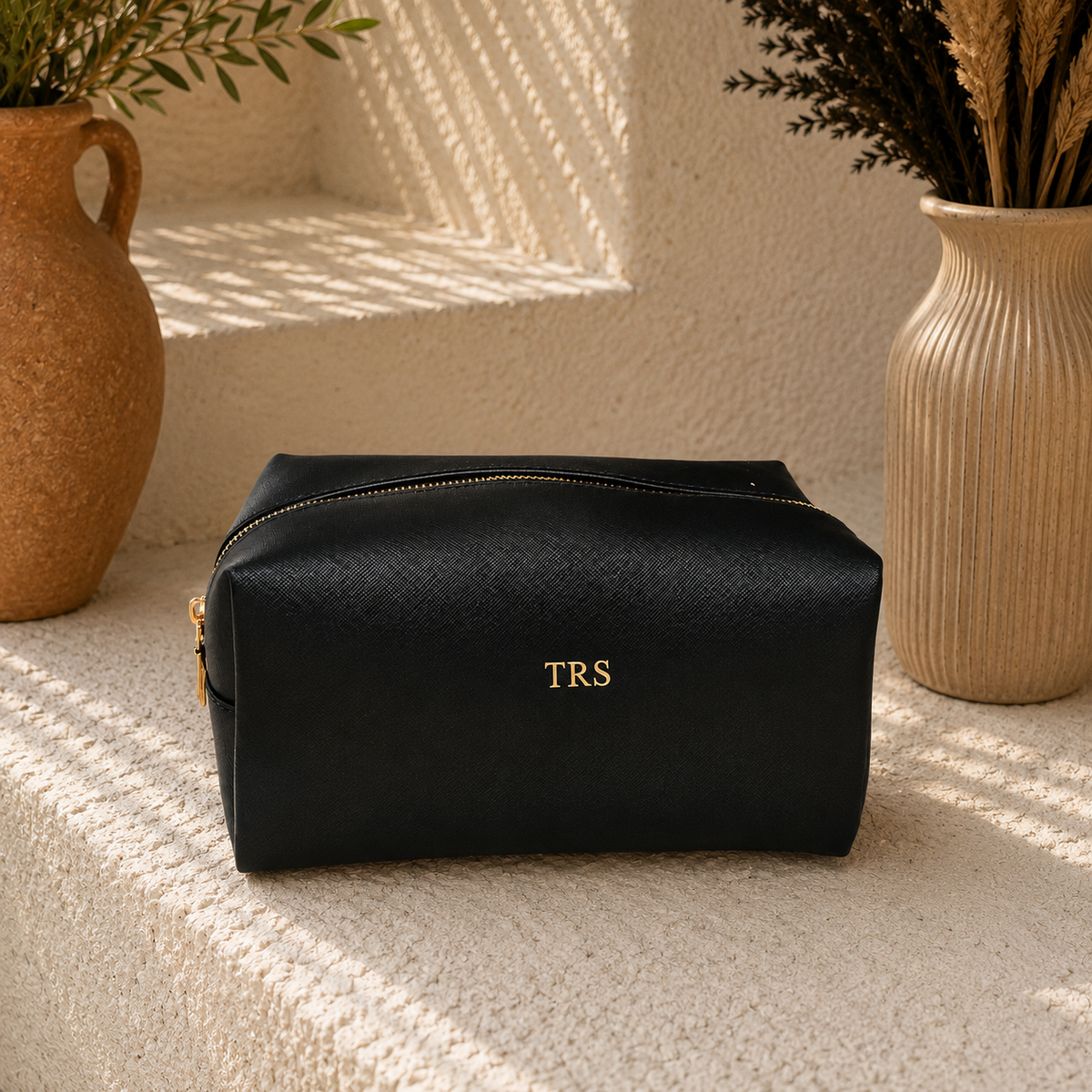 A black textured cosmetic pouch with gold zipper and gold 'TRS' lettering, styled on a textured beige stone surface with decorative vases in the background.