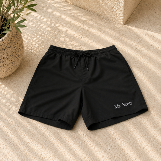 Black drawstring shorts with 'Mr. Scott' embroidered in white on the lower left leg, laid flat on a textured beige surface with a potted plant in the corner.