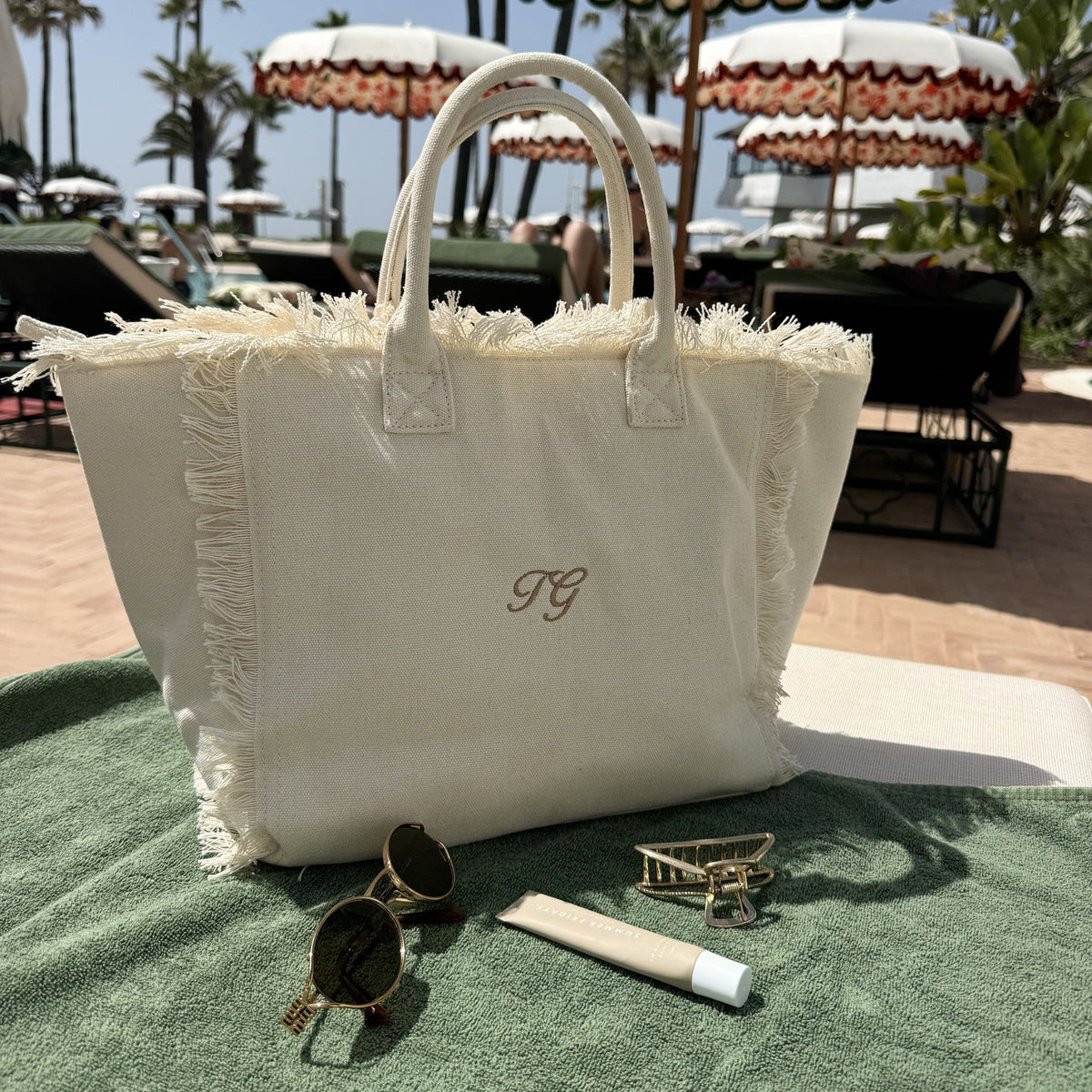 White tote bag with monogram, sunglasses, and lip balm on a green surface with beach umbrellas in the background.