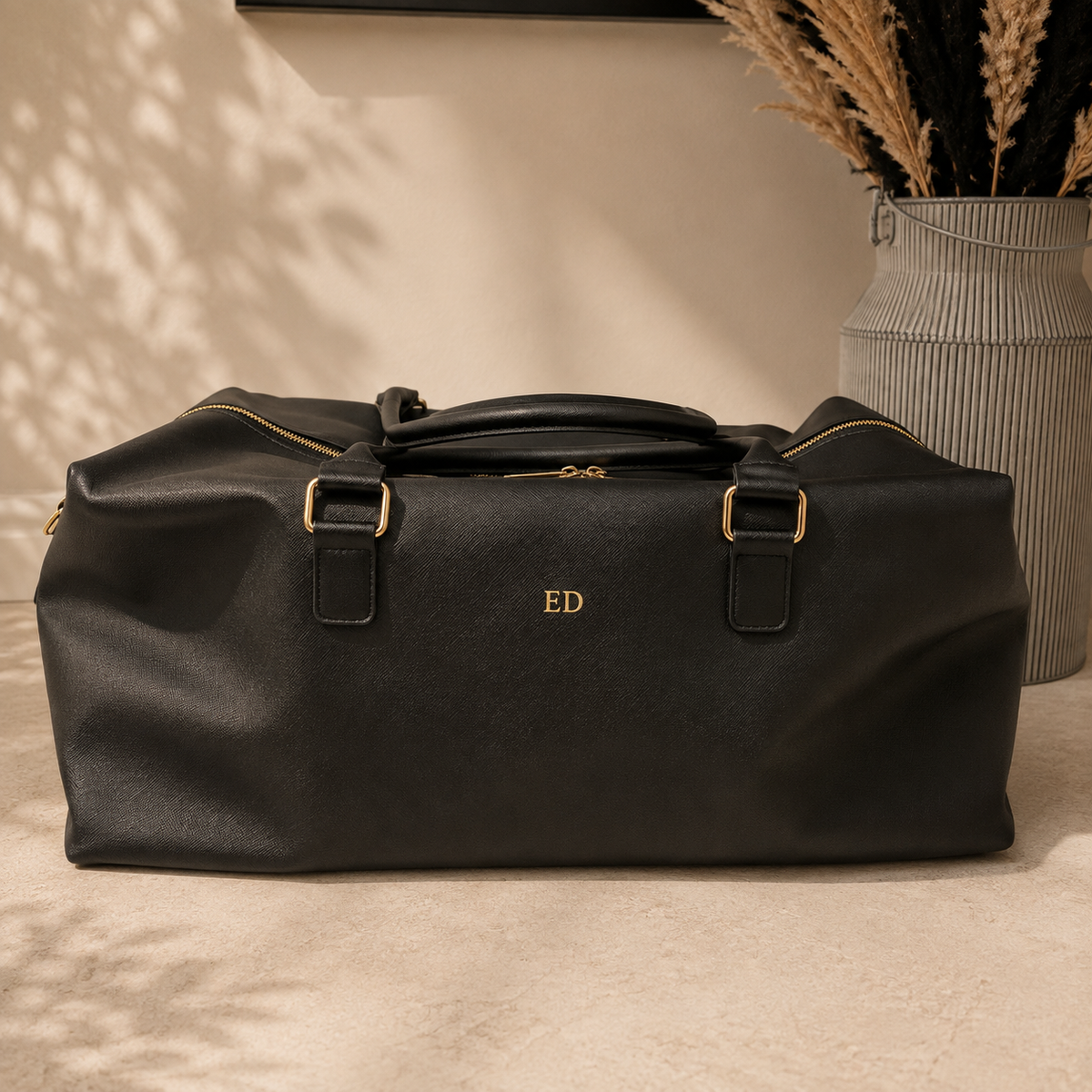 A black textured leather duffel bag with gold hardware and a personalized gold monogram 'ED' on the front, set on a beige floor next to a decorative vase with dried grass.
