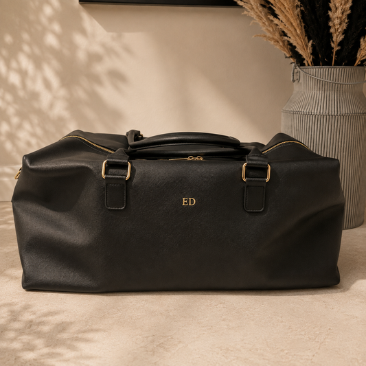 A black textured leather duffel bag with gold hardware and a personalized gold monogram 'ED' on the front, set on a beige floor next to a decorative vase with dried grass.