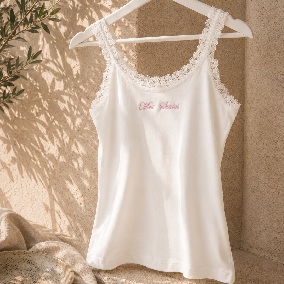 A white sleeveless camisole tank top with delicate lace trim along the scoop neckline and a small pink script logo on the chest, hanging on a white hanger against a textured beige wall with soft leaf shadows.