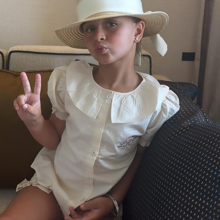 Young girl wearing a white hat and beige shorts set with personalisation sitting on a couch.