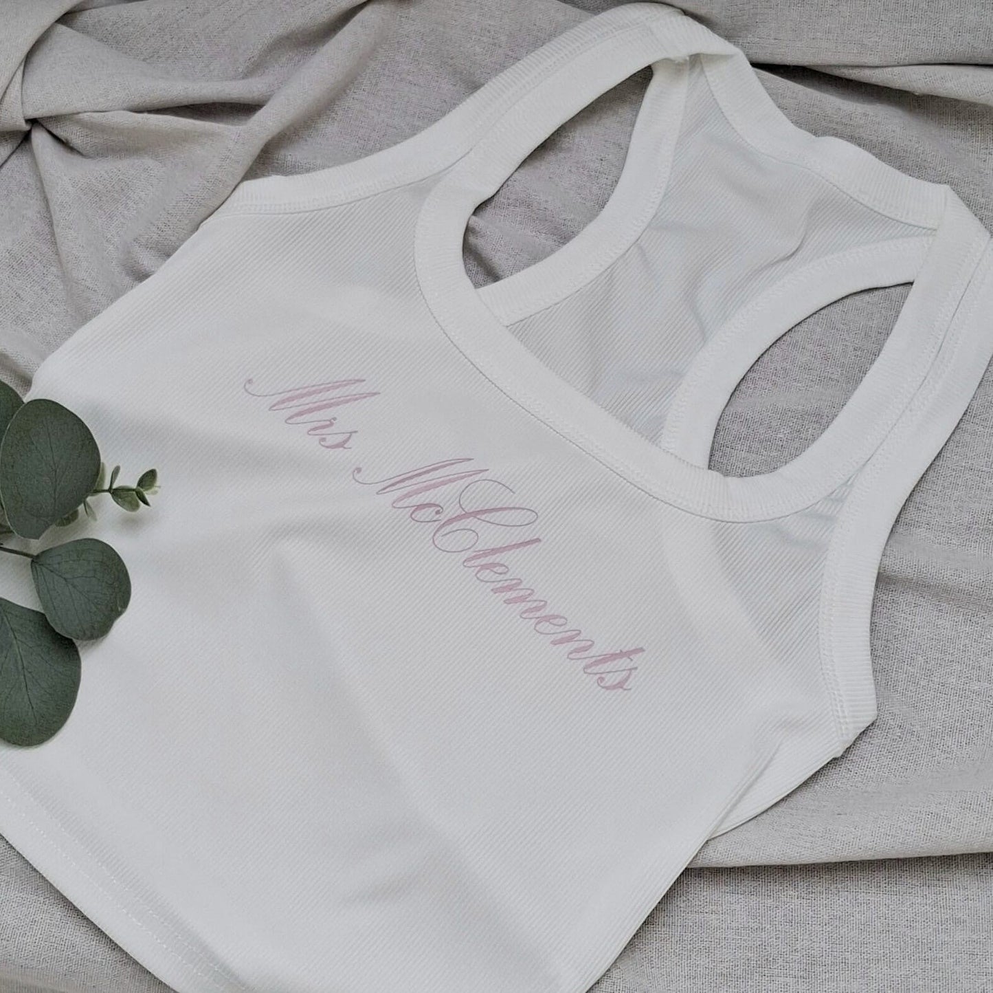 White tank top with cursive text on a gray surface with green leaves.
