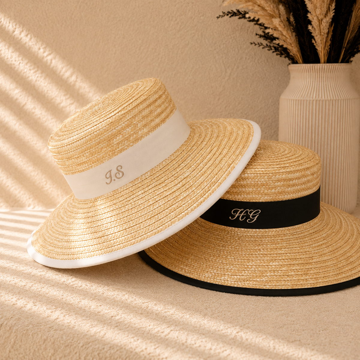 Two woven straw sun hats with wide brims are arranged on a beige textured surface. One hat has a white band with a gold monogram, and the other has a black band with a gold monogram.