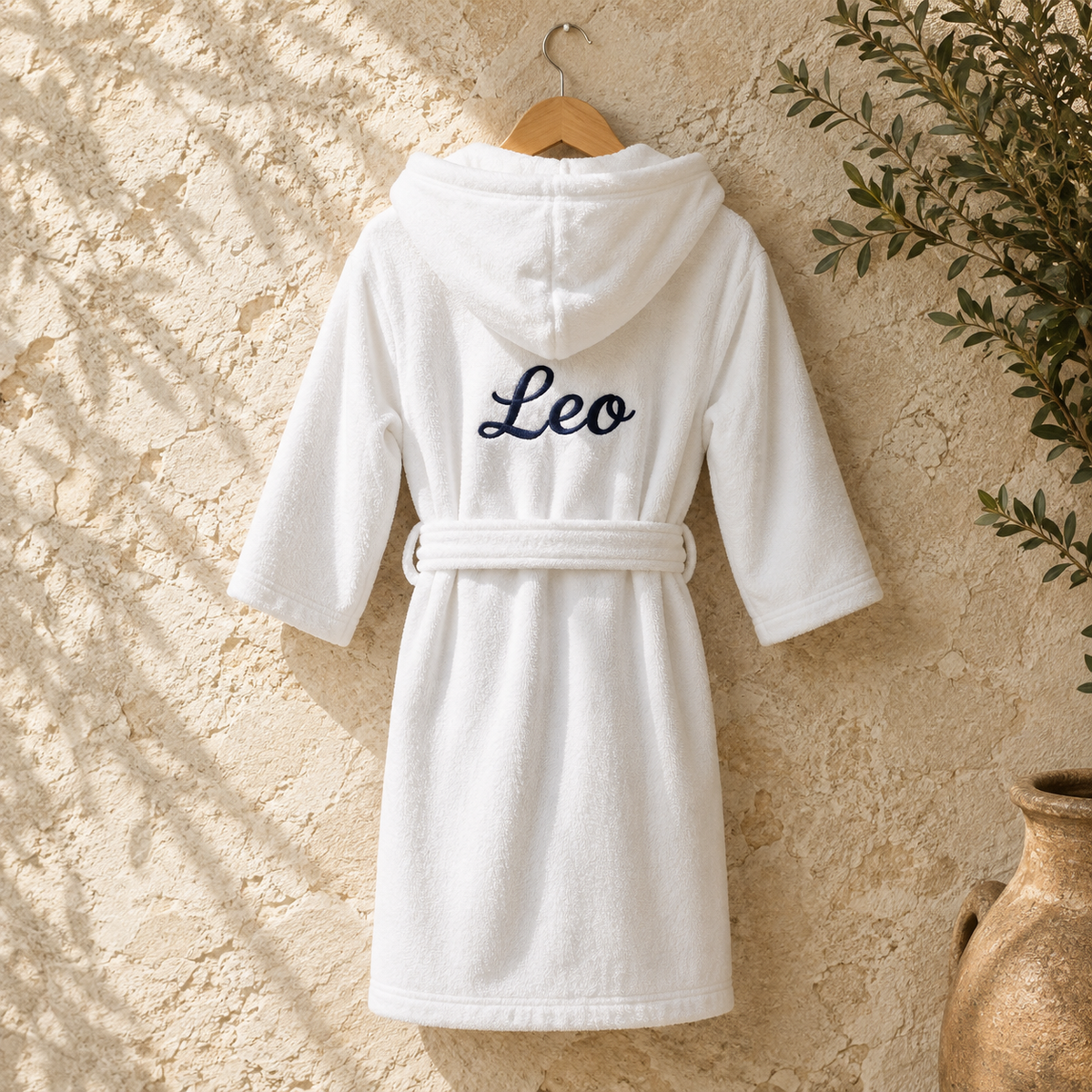 A white hooded bathrobe with a matching belt hangs on a wooden hanger against a textured beige wall. The back of the robe is embroidered with the name 'Leo' in dark navy script.
